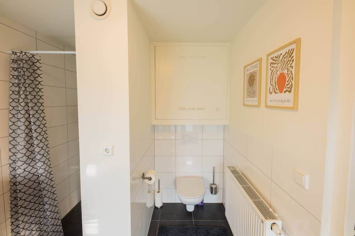 A bathroom with a white toilet, a shower with a fish-scale pattern, and two framed botanical artwork on the wall.