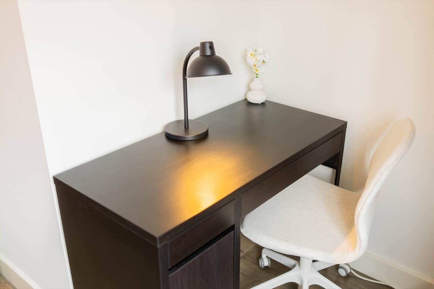 Un bureau en bois foncé, sur lequel se trouve une lampe, une chaise blanche et un petit vase avec des fleurs.