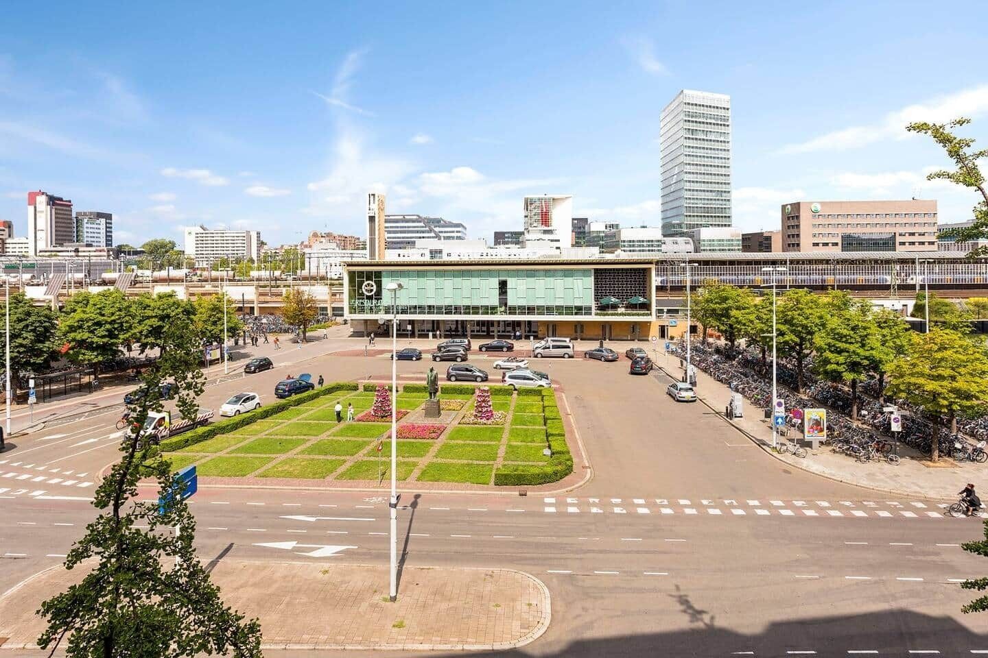 En moderne bygning med en glasfacade, omgivet af et anlagt område med træer og en parkeringsplads.