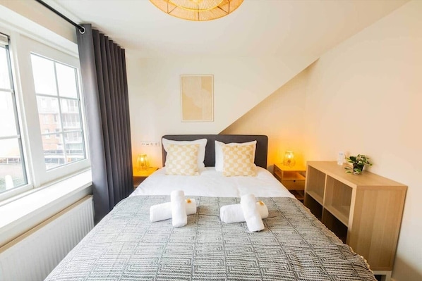 Habitación de hotel con cama, lámparas de noche, mesita de noche, ventana con cortinas y vistas a edificios.