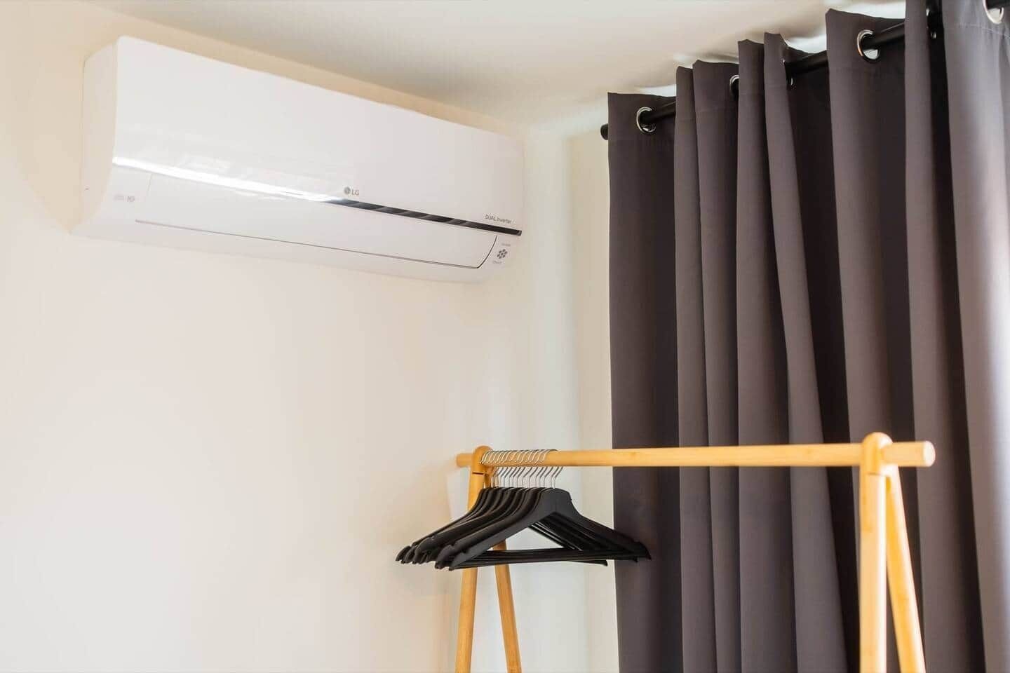 A room with a wall-mounted air conditioner, a wooden hanger rack with clothes, and a set of dark curtains.