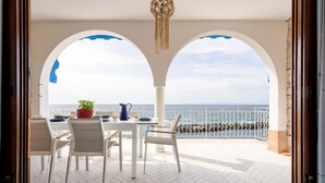 Une terrasse avec une table dressée pour le repas, des chaises blanches et une vue sur la mer, visible à travers des ouvertures en arc de cercle.