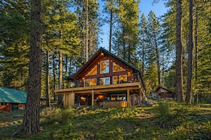 A two-story log cabin with a balcony, surrounded by tall pine trees. - Riverhouse 3 Bedroom Home (Leavenworth)