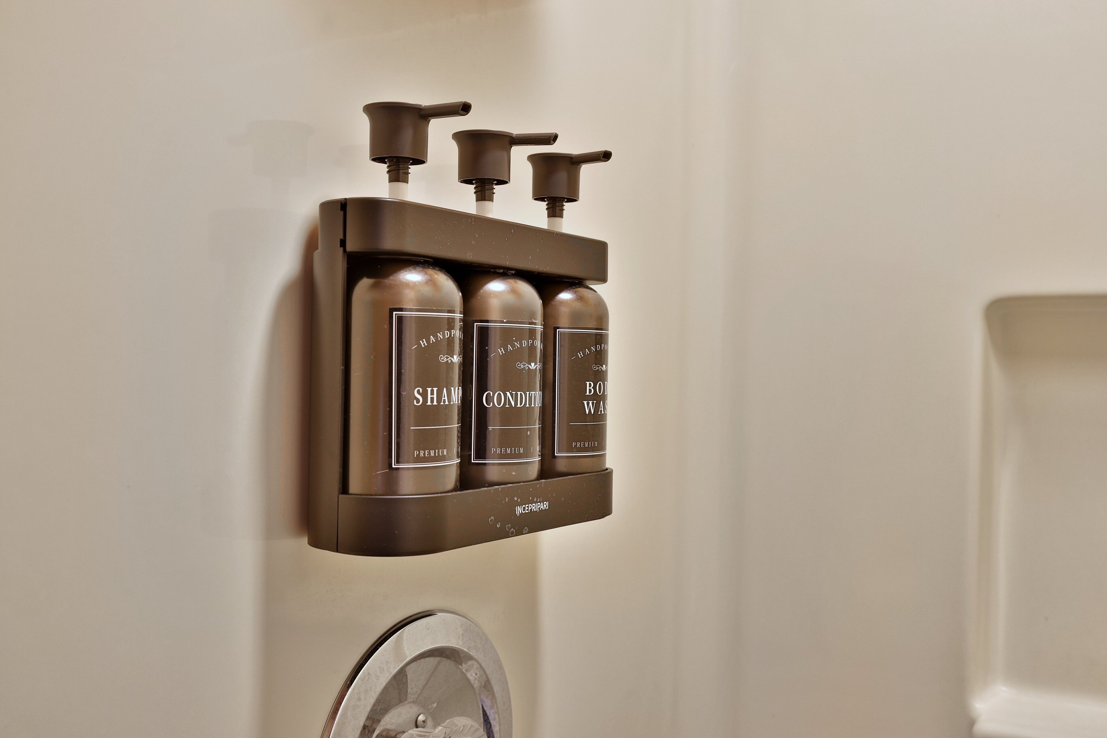 Un dispenser a parete con tre flaconi etichettati 'Shampoo', 'Balsamo' e 'Detergente corpo'.
