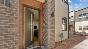 A modern building entrance with a brick facade, a wooden door, and a small garden area with shrubs and a metal sculpture. - Villa #24 At Bloomington Country Club 4 Bedroom Townhouse (St. George)