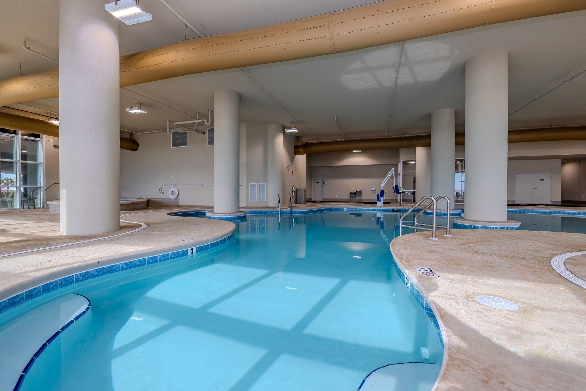 Ein Hallenbad mit klarem, blauem Wasser, umgeben von beigen Fliesen und weißen Säulen.