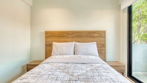 A bedroom with a wooden headboard, a bed with a patterned bedspread, two bedside tables, and a window with a view of trees.
