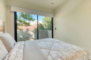 A bedroom with a large bed, a sliding glass door leading to a balcony, and a view of buildings and trees outside.