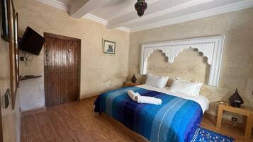 A bedroom with a wooden door, a bed with a blue and white striped bedspread, a white headboard with intricate carvings, and a small wooden nightstand with a lamp.