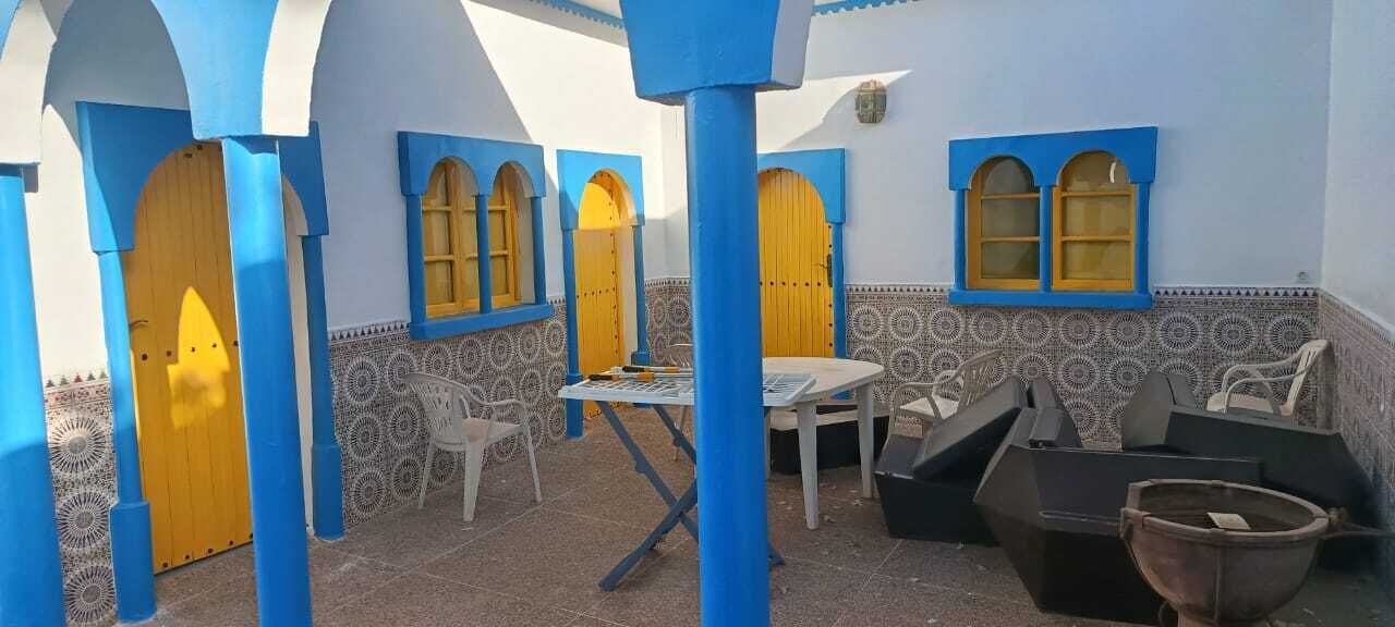 Um pátio com portas azuis e amarelas, uma mesa, cadeiras e uma lixeira preta.