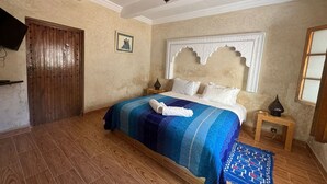 A bedroom with a wooden door, a bed with a blue and white striped bedspread, a decorative headboard, and a small wooden nightstand with a lamp. - LES ETOILES DU KSAR (Mirleft)