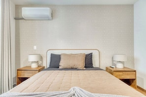 A hotel room with a bed, two bedside tables, a wall-mounted air conditioner, and a patterned wall.