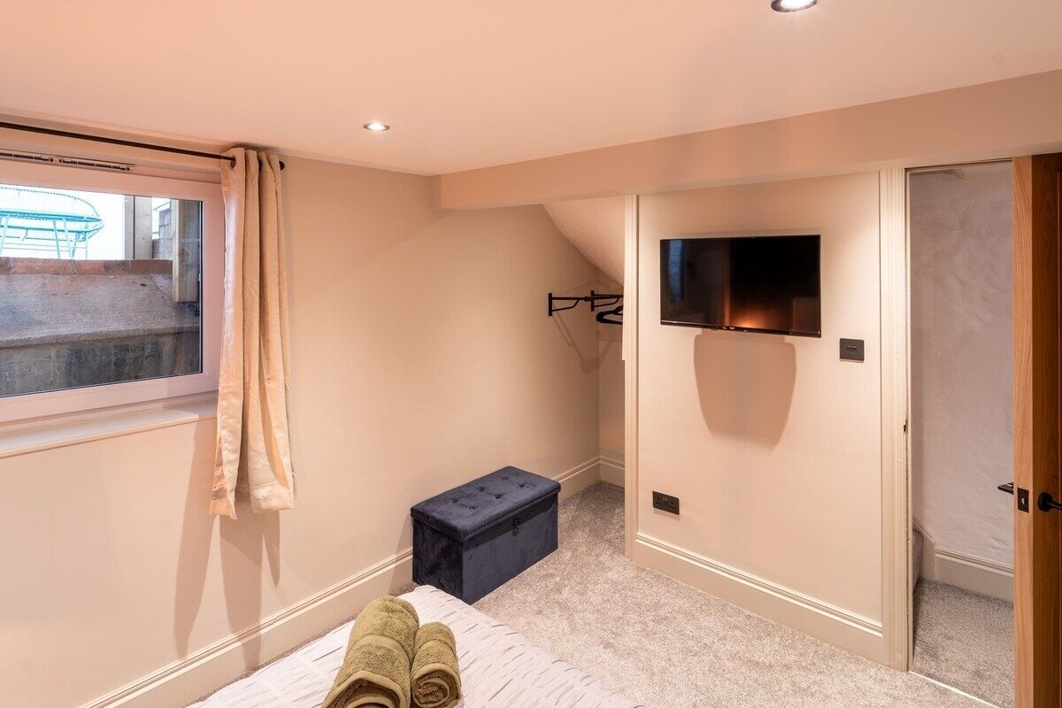 A bedroom with a bed, a TV mounted on the wall, a small storage box, and a window with curtains.