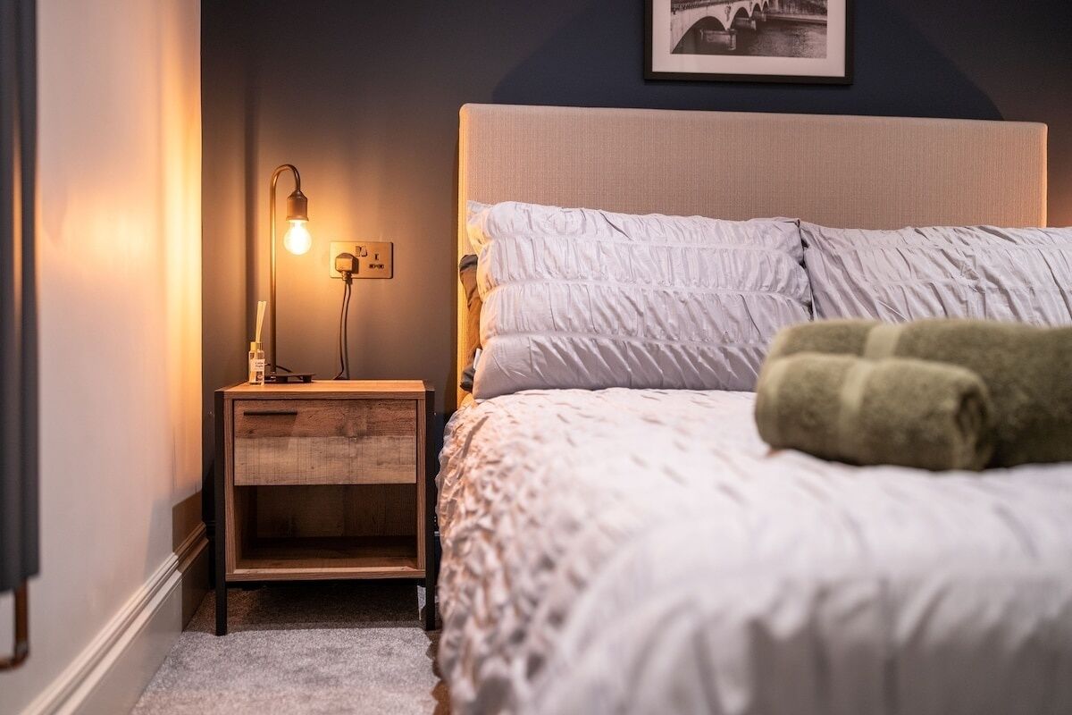 En pænt redt seng med hvid dyne og puder, en sengetårnslampe og et billede i ramme på væggen.