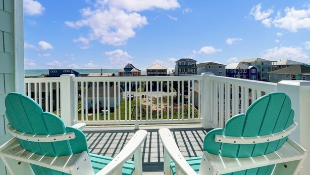 Un balcon agrémenté de deux fauteuils Adirondack turquoise, d’une balustrade blanche et offrant une vue sur l’océan et les maisons de plage.