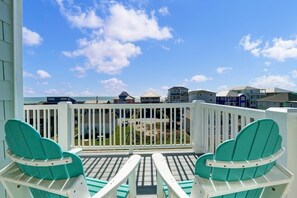 Un balcon agrémenté de deux fauteuils Adirondack turquoise, d’une balustrade blanche et offrant une vue sur l’océan et les maisons de plage.