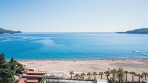 A beach with clear blue water, a sandy shore, and a few buildings in the foreground. - Hotel Invictus (Becici)