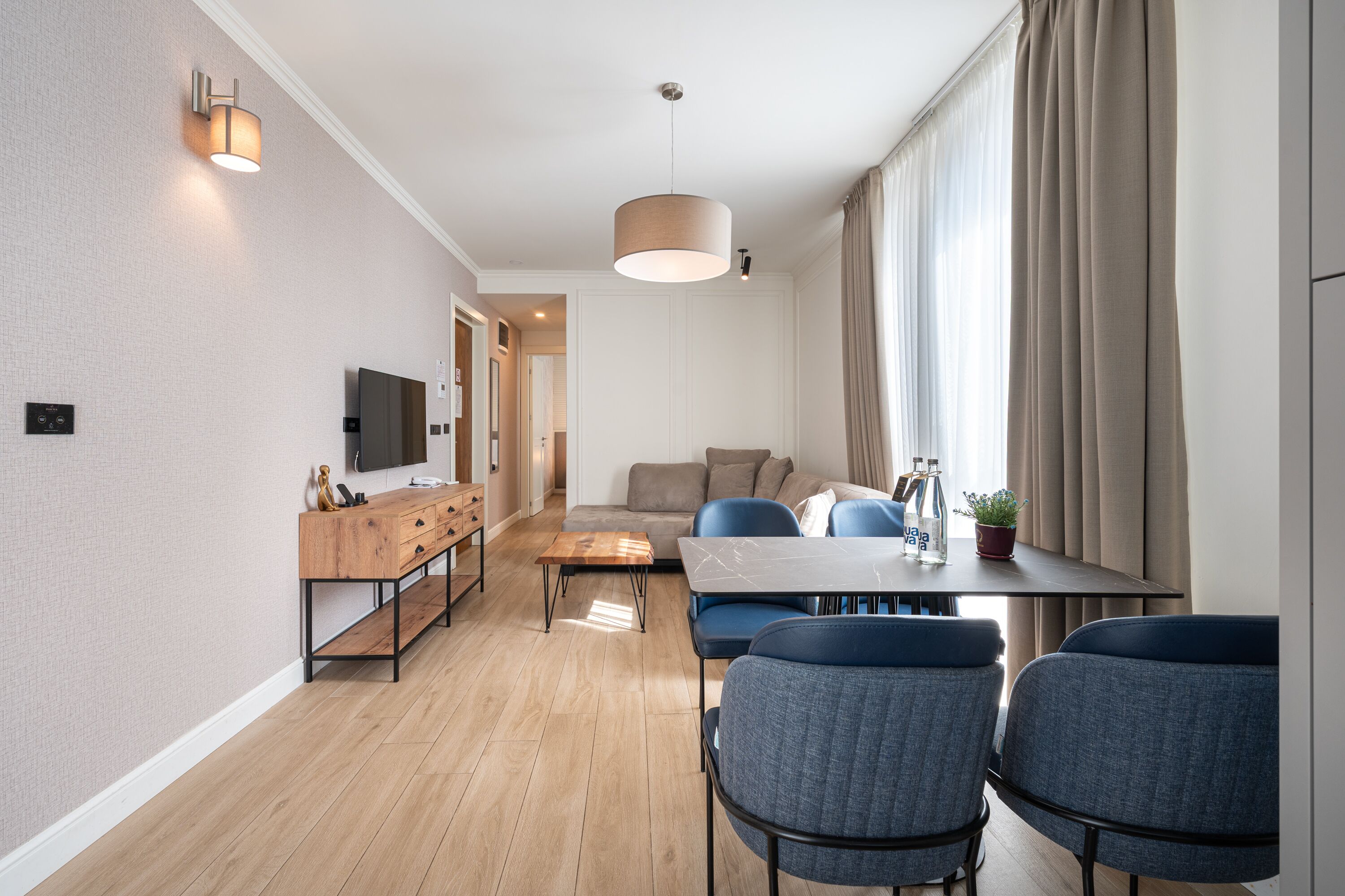 A modern living room with a sofa, a dining table with chairs, a wooden coffee table, and a TV mounted on the wall.