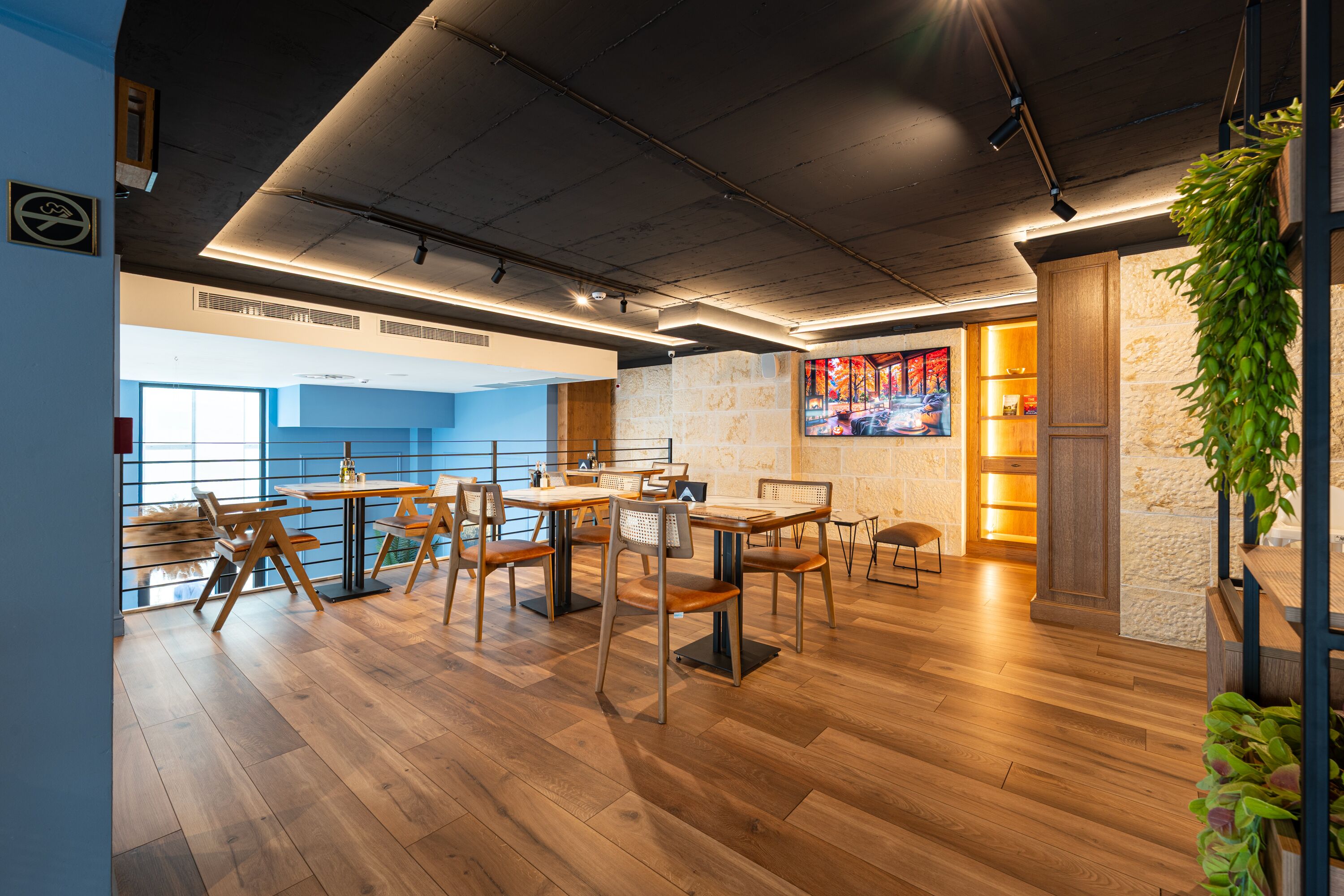A modern restaurant interior with wooden flooring, multiple tables and chairs, and a large window providing natural light.