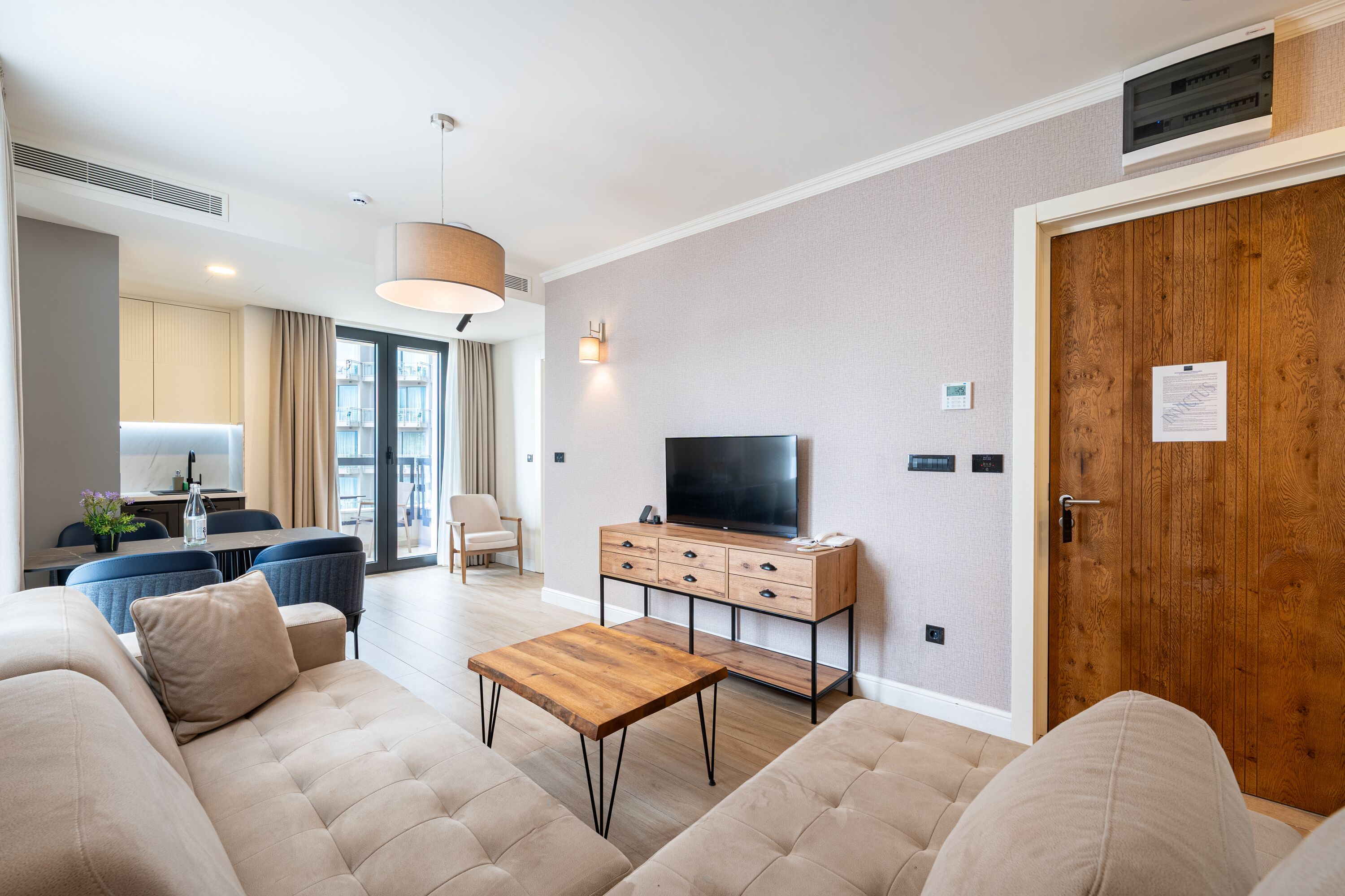 Ein modernes Wohnzimmer mit einer Couch, einem Holztisch, einem Fernseher auf einem Holzständer und einem Essbereich im Hintergrund.
