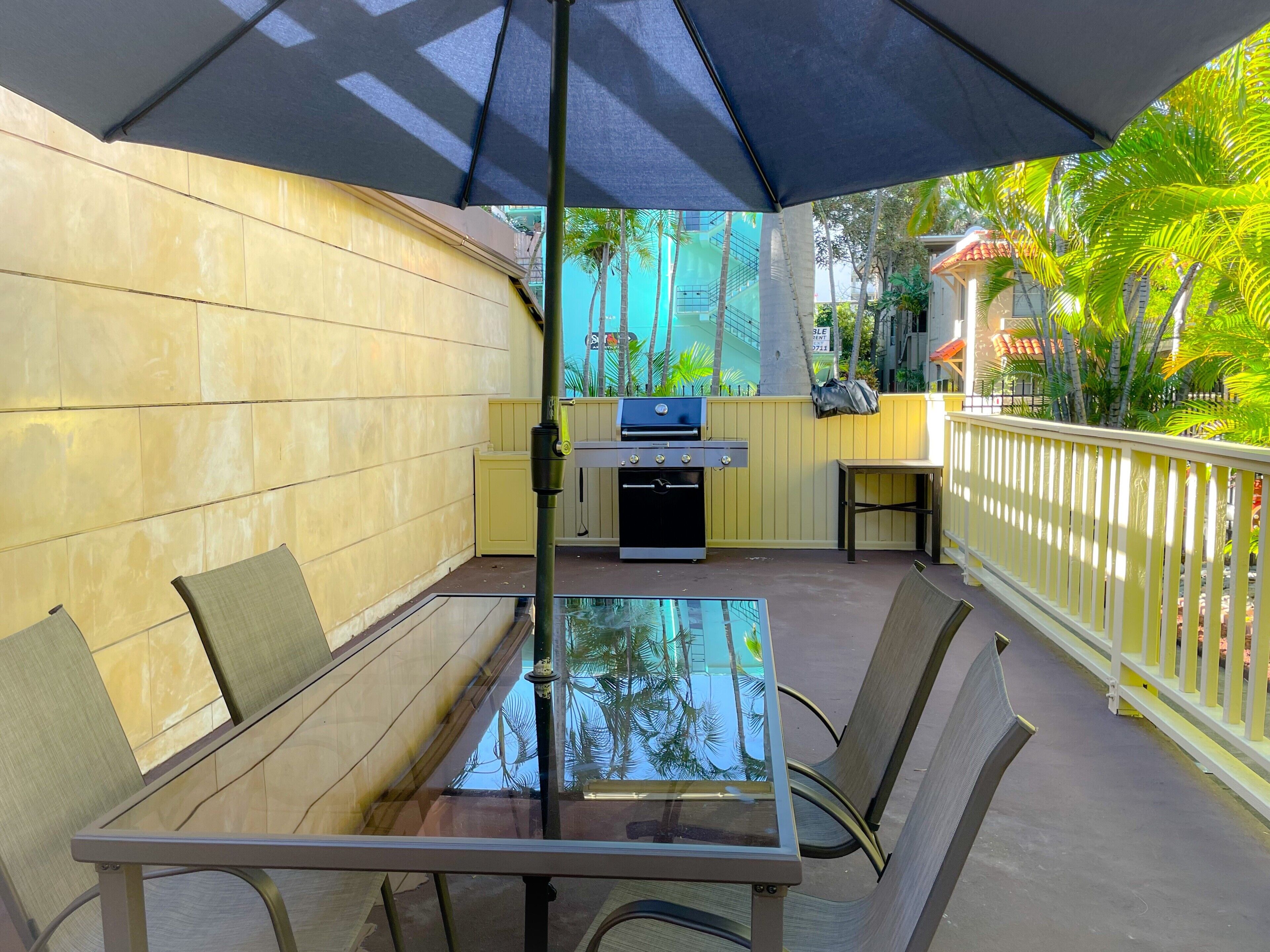 Une terrasse extérieure avec une table et des chaises en verre, un barbecue et une clôture jaune.