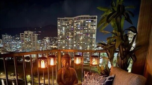 A balcony with a Buddha statue, wicker furniture, and city lights in the background. - Honu Hideaway Studio at Royal Garden | Lanai Oasis (Honolulu)