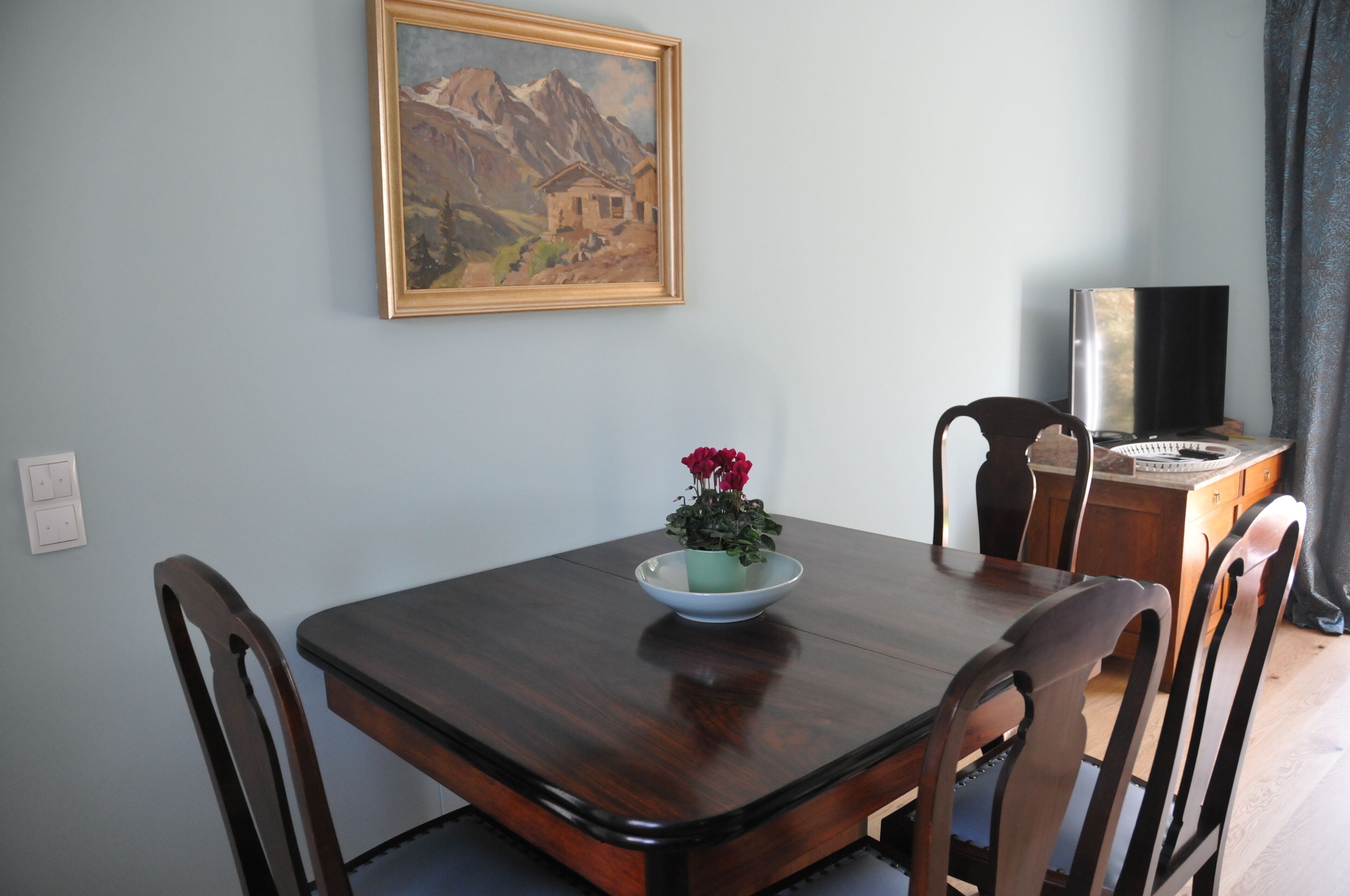 Un comedor con mesa de madera, cuatro sillas, un cuadro en la pared y una pequeña maceta con flores rojas.