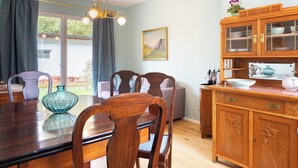 A dining area with a wooden table and chairs, a kitchen with wooden cabinets and a countertop, and a large window with curtains. - Penta Apartments (Gamlitz)