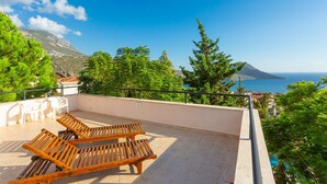 A balcony with two wooden lounge chairs, a white chair, and a view of the sea and mountains.