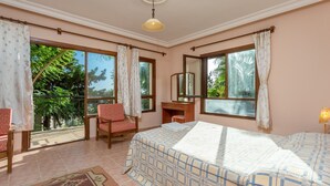 A bedroom with a bed, a desk, a chair, and a balcony with a view of trees.