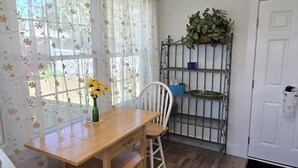 Un comedor con una mesa de madera y dos sillas blancas, una ventana con cortinas florales y un estante metálico con plantas.