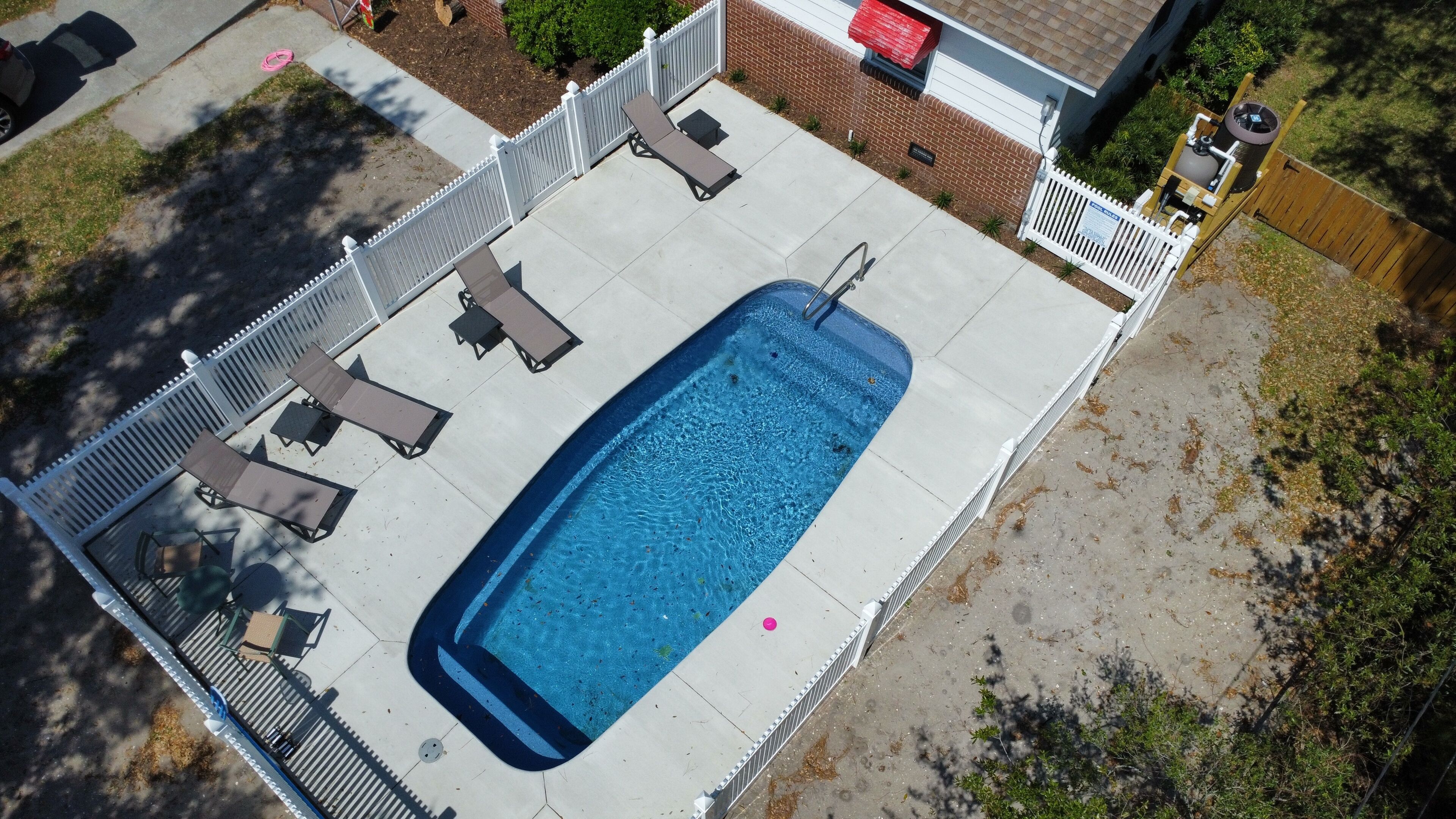 Vue aérienne d’une piscine rectangulaire bordée d’une clôture blanche, entourée de chaises longues et d’un espace patio couvert.