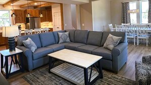 A modern living room with a sectional sofa, a coffee table, and a dining area with white chairs. - DC51: Walter's Watch - Brand New Construction (Ocracoke)