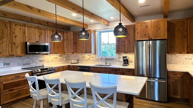 A modern kitchen with wooden cabinets, a stainless steel refrigerator, a large island with bar stools, and pendant lighting. - DC51: Walter's Watch - Brand New Construction (Ocracoke)