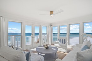 A spacious room with a large window overlooking a beach, a ceiling fan, and a round table with a bowl.