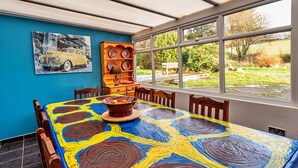 Een eetruimte met een tafel bedekt met een kleurrijk abstract schilderij, een houten buffetkast met borden, en uitzicht op een tuin door grote ramen.
