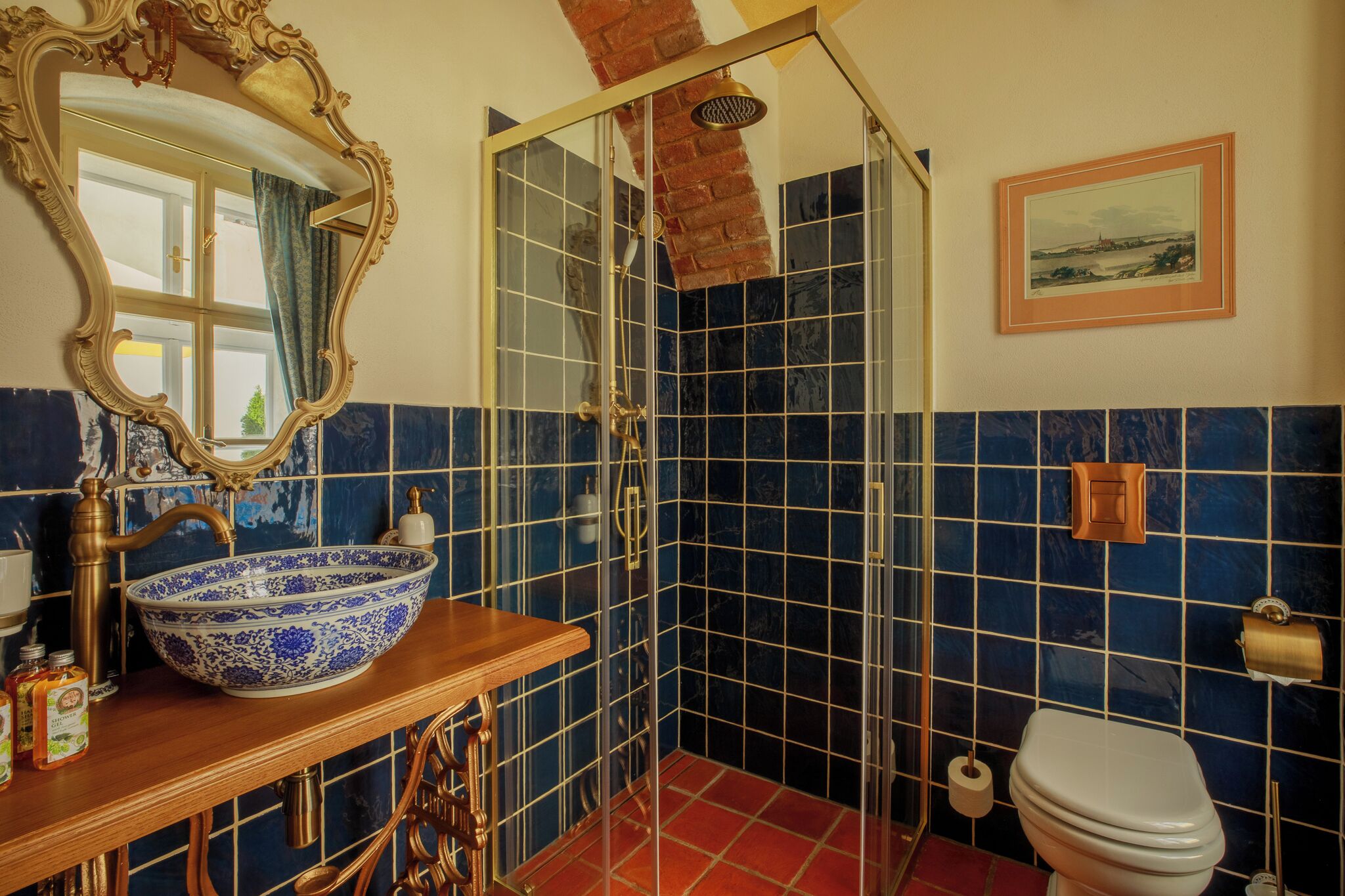 A bathroom with a large blue and white ceramic sink, a wooden vanity, a glass-enclosed shower, a toilet, and a framed picture on the wall.