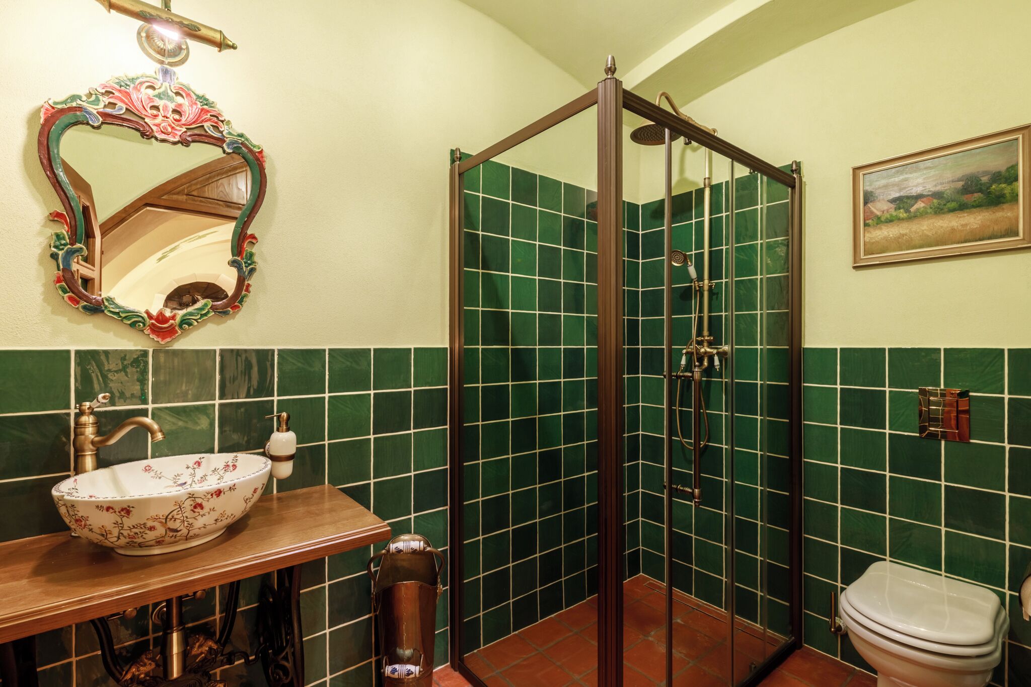 Un baño con tocador de madera, un lavamanos blanco con motivos florales, un box de ducha con azulejos verdes y un inodoro blanco.