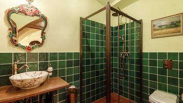 Un baño con tocador de madera, un lavamanos blanco con motivos florales, un box de ducha con azulejos verdes y un inodoro blanco.