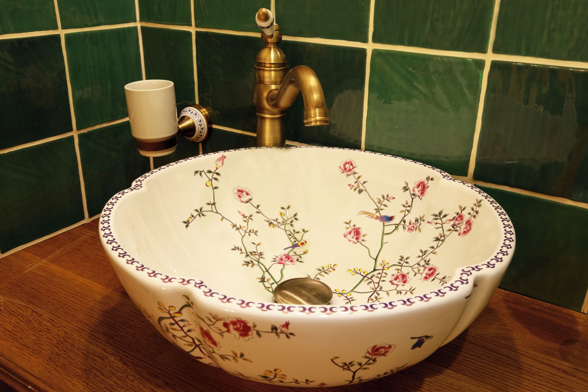 A white ceramic sink with floral patterns, a brass faucet, and a white cup on the wall.
