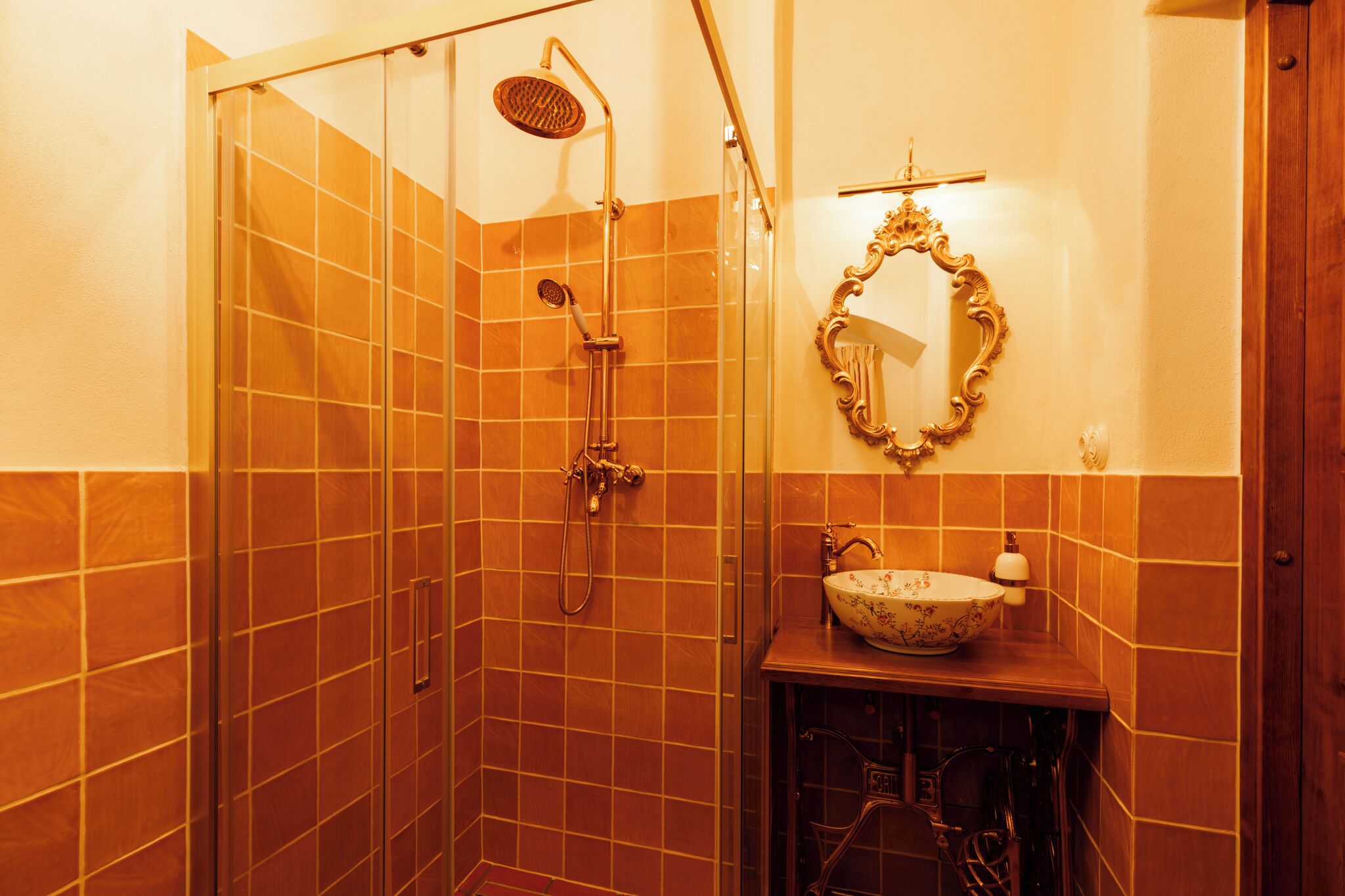 Un baño con ducha envuelta en vidrio, tocador de madera con lavabo y un espejo decorativo en la pared.
