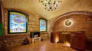 Una habitación con paredes de piedra, dos grandes ventanales de vidrio emplomado, una chimenea y barriles de madera.