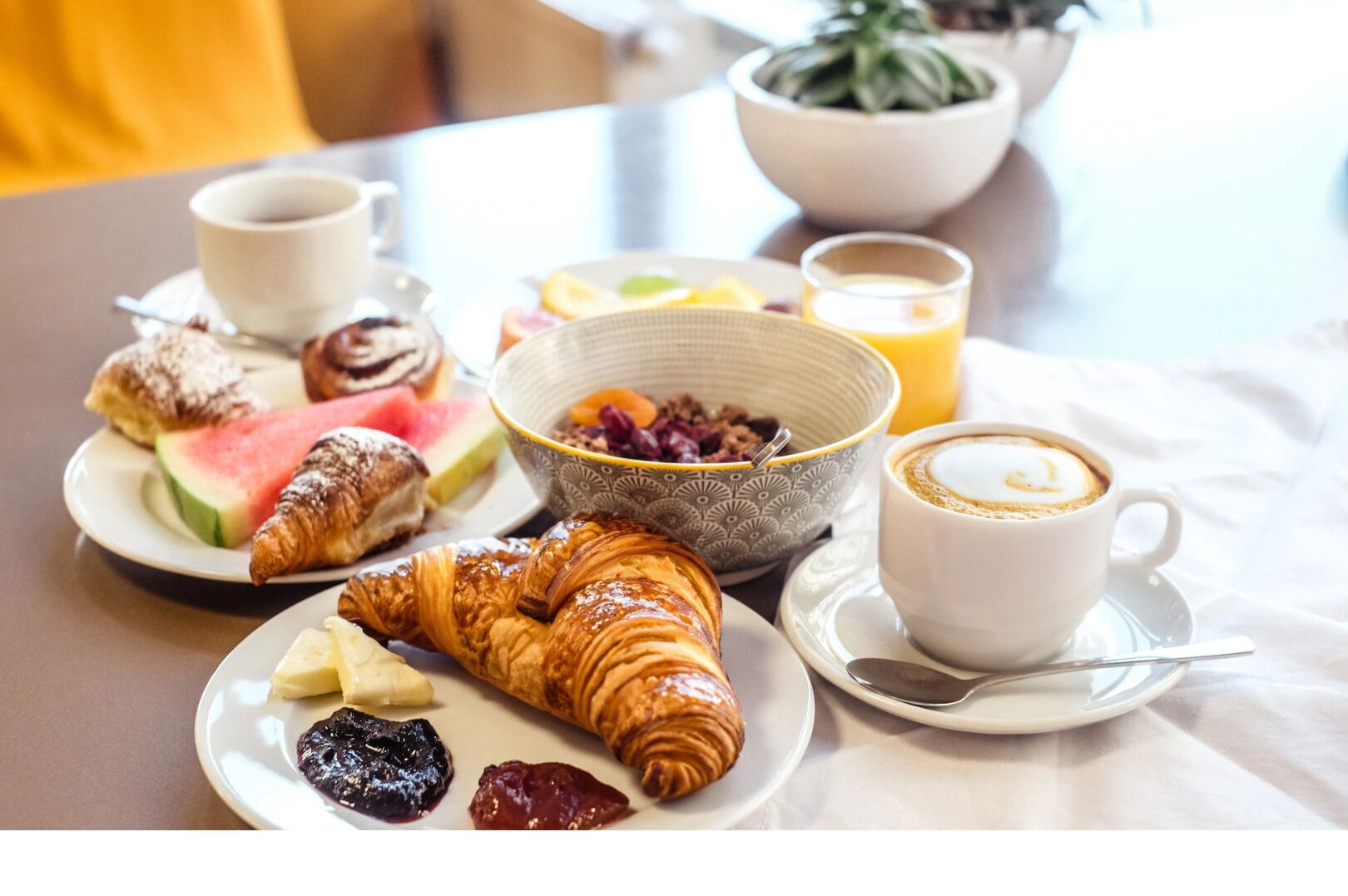Een ontbijttafel met croissants, fruit, een kom ontbijtgranen, een kop koffie en een glas sinaasappelsap.