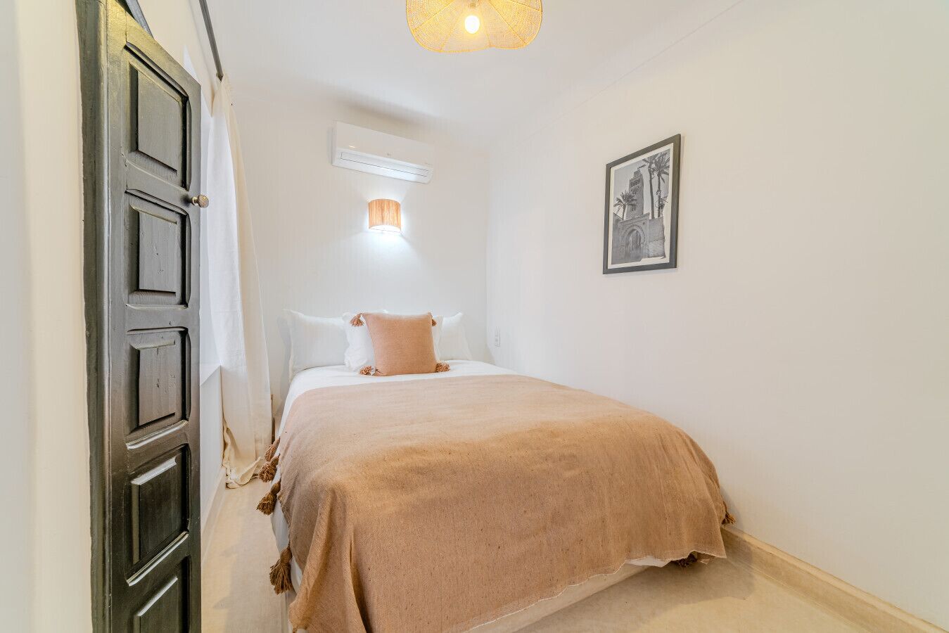 Un couloir étroit mène à une chambre où se trouvent un lit, un climatiseur fixé au mur et un tableau encadré.