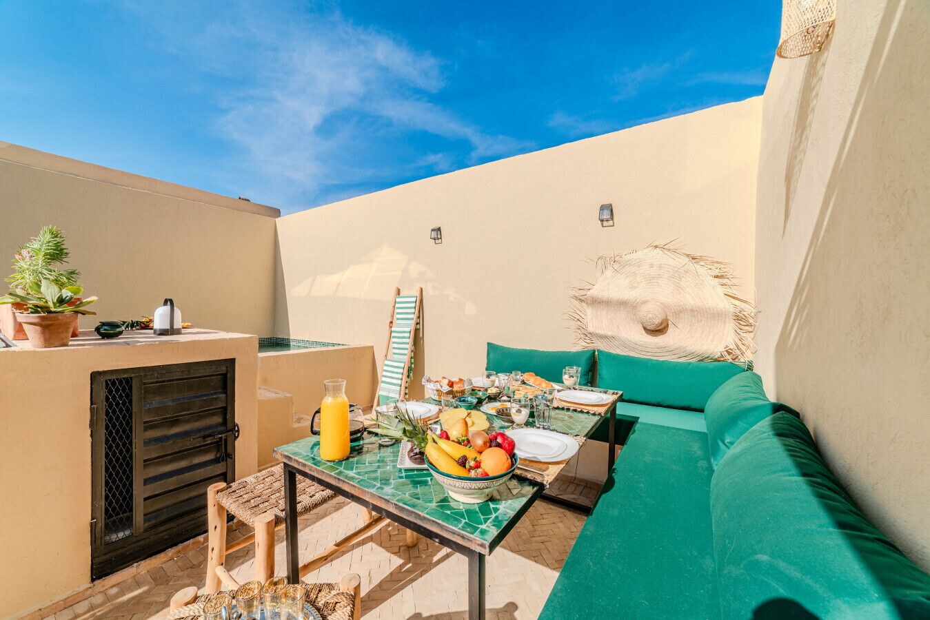 A small outdoor dining area with a table set for two, a green cushioned bench, a potted plant, and a wall-mounted light fixture.