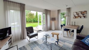 A modern living room with a TV, sofa, dining table, and chairs. There is a large window with curtains, a lamp, and a painting on the wall. - Summio Villapark Schildmeer (Steendam)