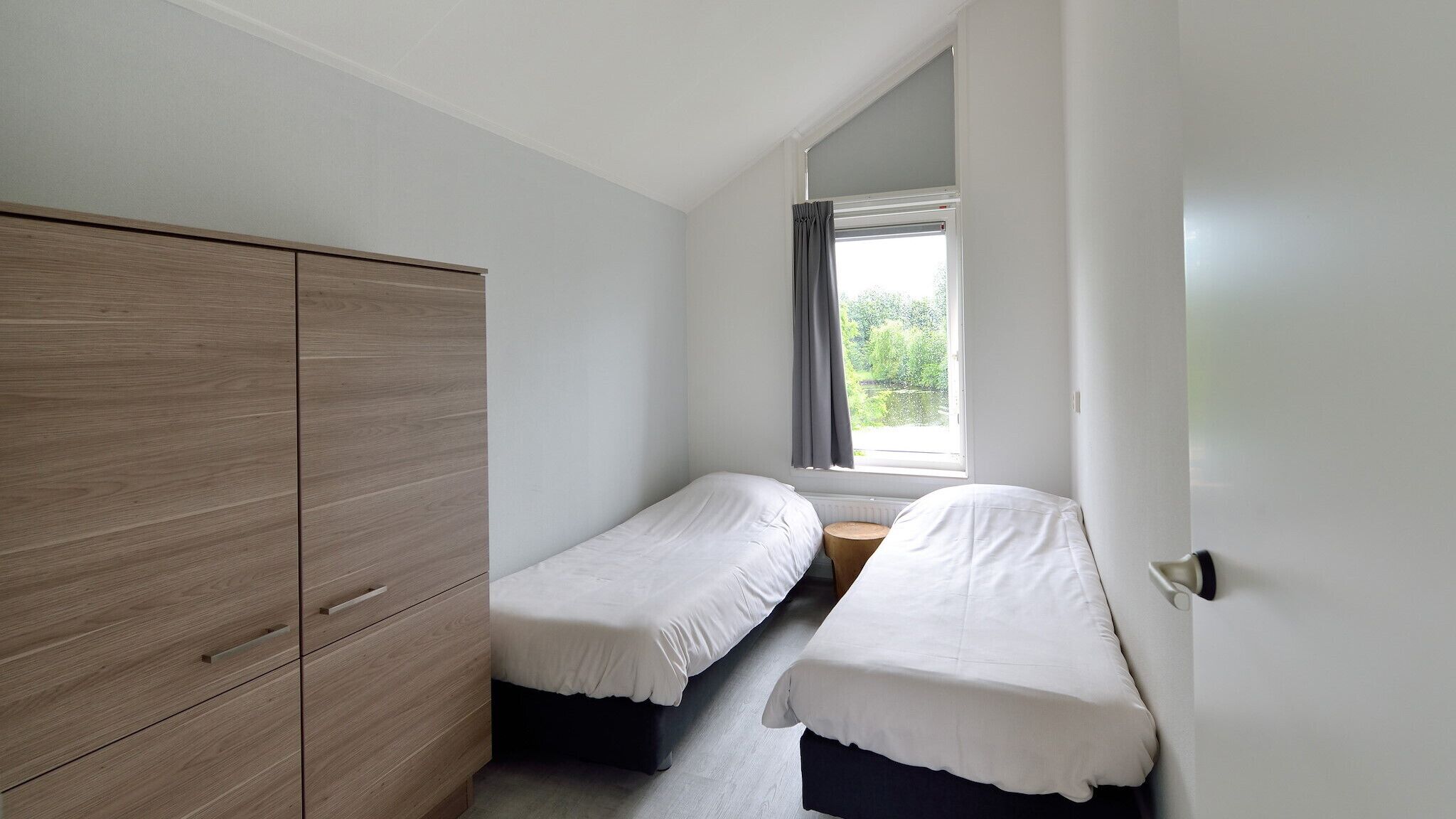 A small, clean room with two single beds, a wooden wardrobe, and a window with a view of greenery.