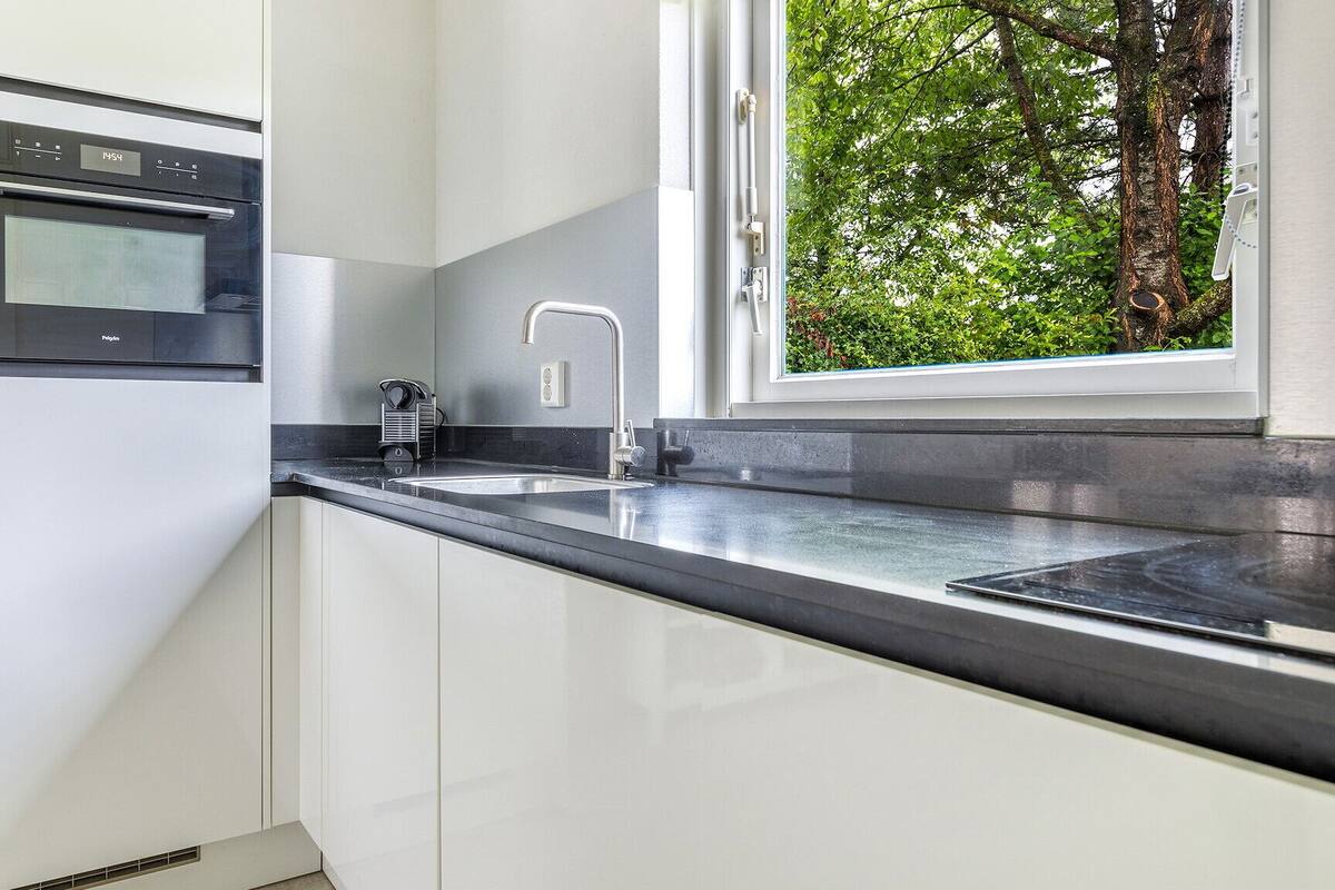A modern kitchen with a built-in oven, a stainless steel sink, and a window overlooking greenery.