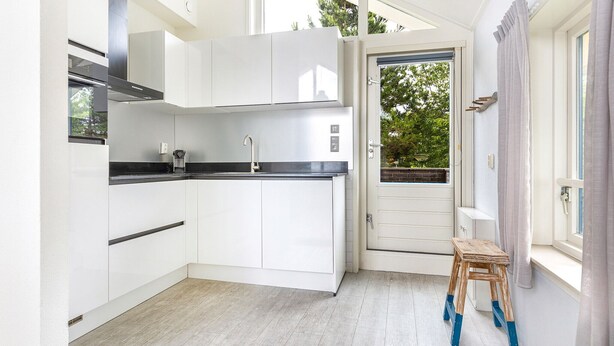 A modern kitchen with white cabinets, a black countertop, and a built-in stove. There's a small wooden stool and a window with curtains. - Summio Villapark Schildmeer (Steendam)