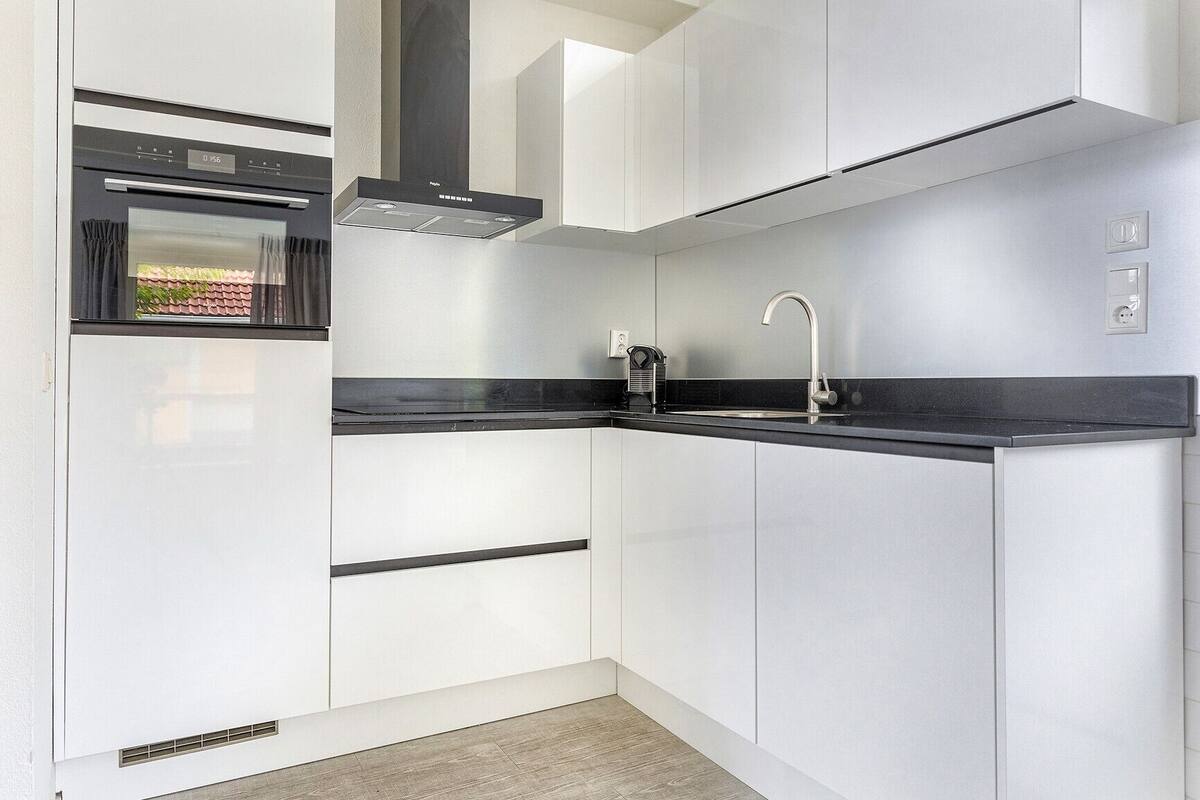 A modern kitchen with white cabinets, a black countertop, a built-in oven, a stainless steel range hood, and a sink with a faucet.